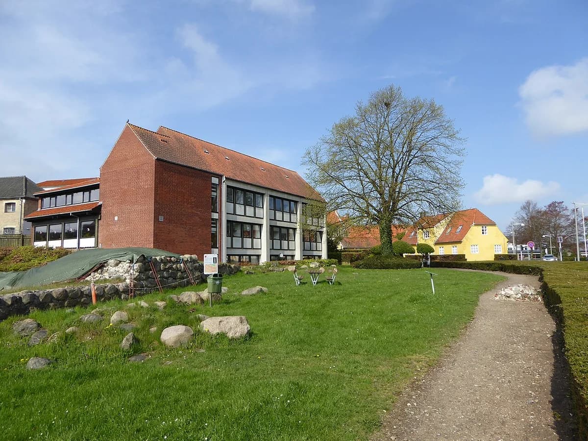 A grassy area with stones, a tree, and modern buildings under a blue sky.
