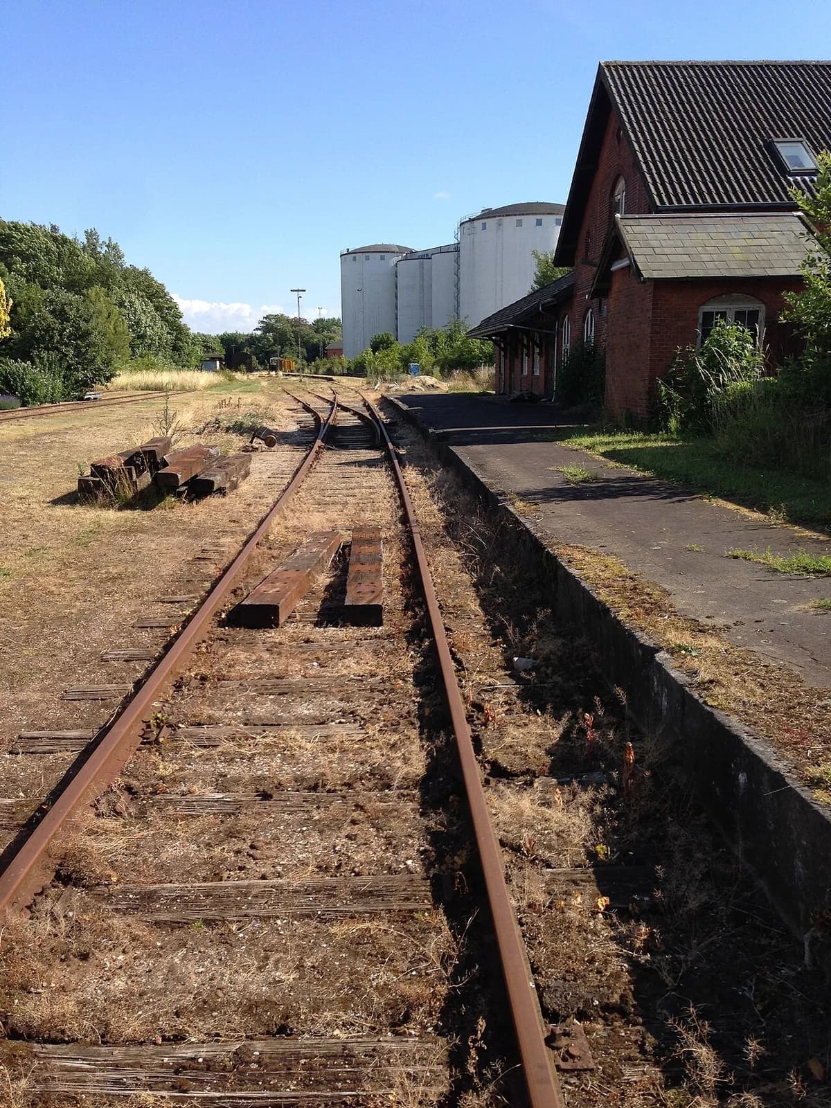 Railway tracks beside a brick station building with silos in the distance.