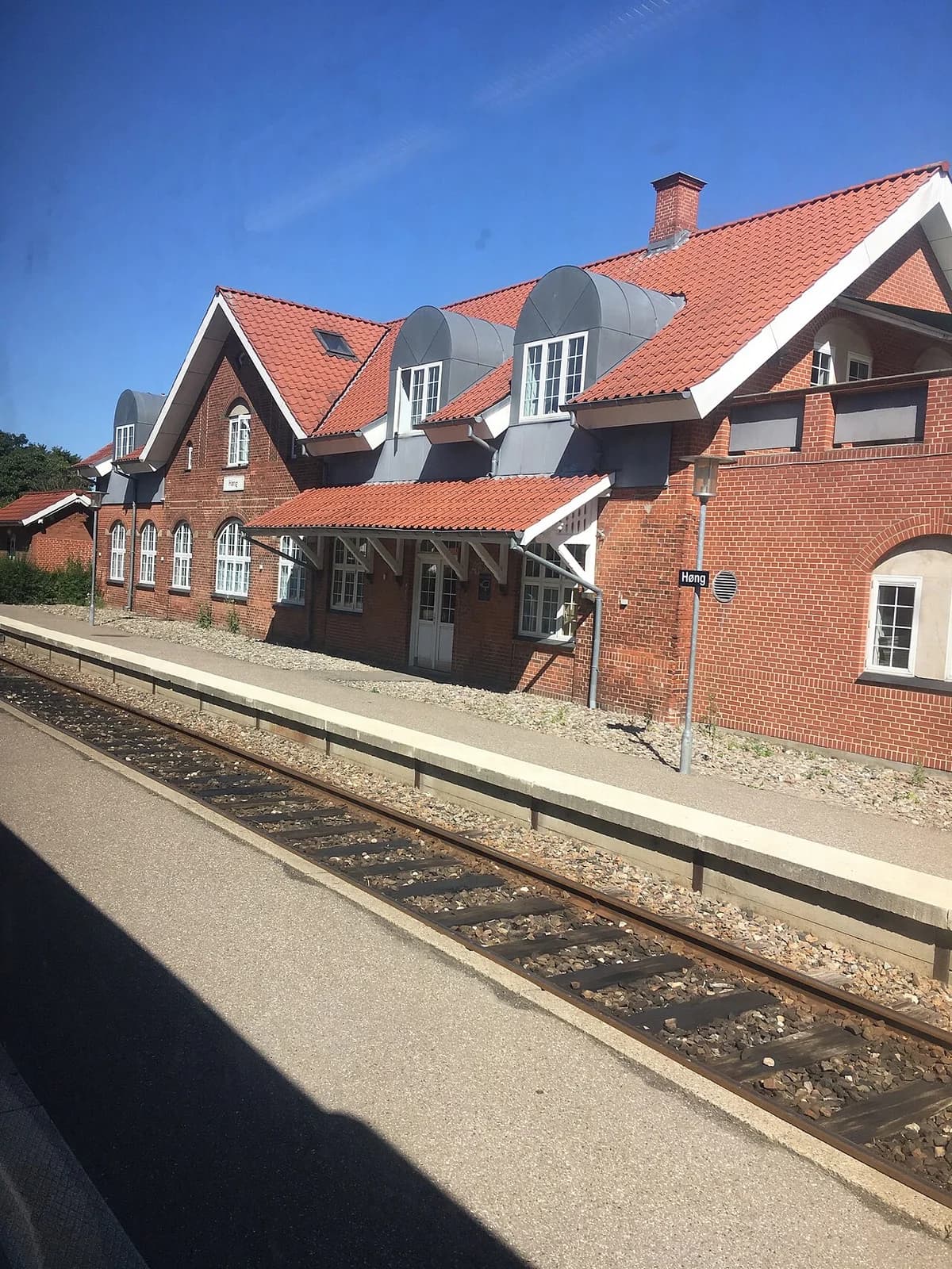 Red-brick railway station building beside tracks and platform under a clear blue sky.
